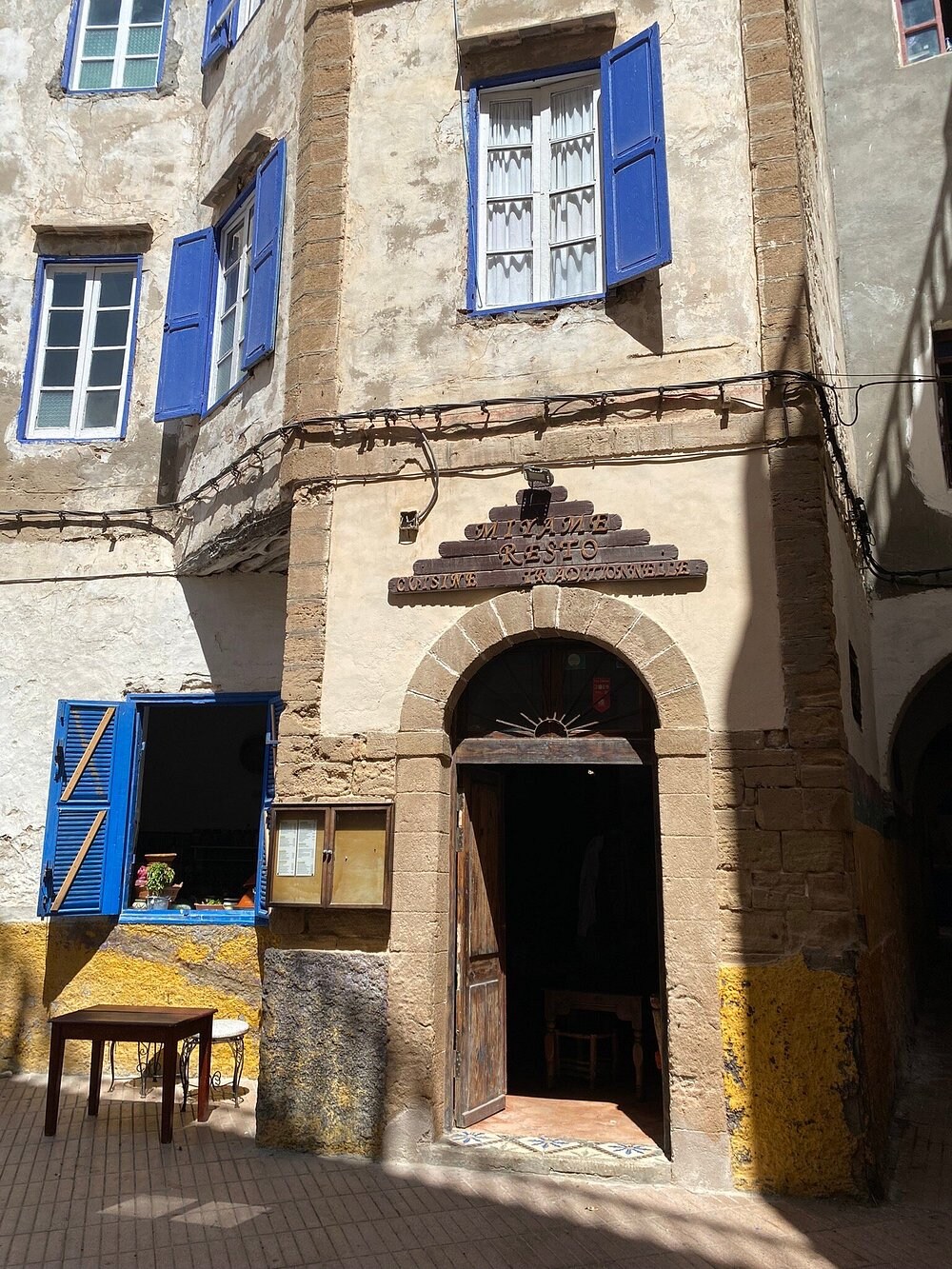 An exterior view of a historic building features earthy stone walls with a weathered texture. Blue shutters frame a large window above a welcoming arched door. A small table is positioned outside, and nearby, the vibrant colors of the well-trodden ground provide contrast.