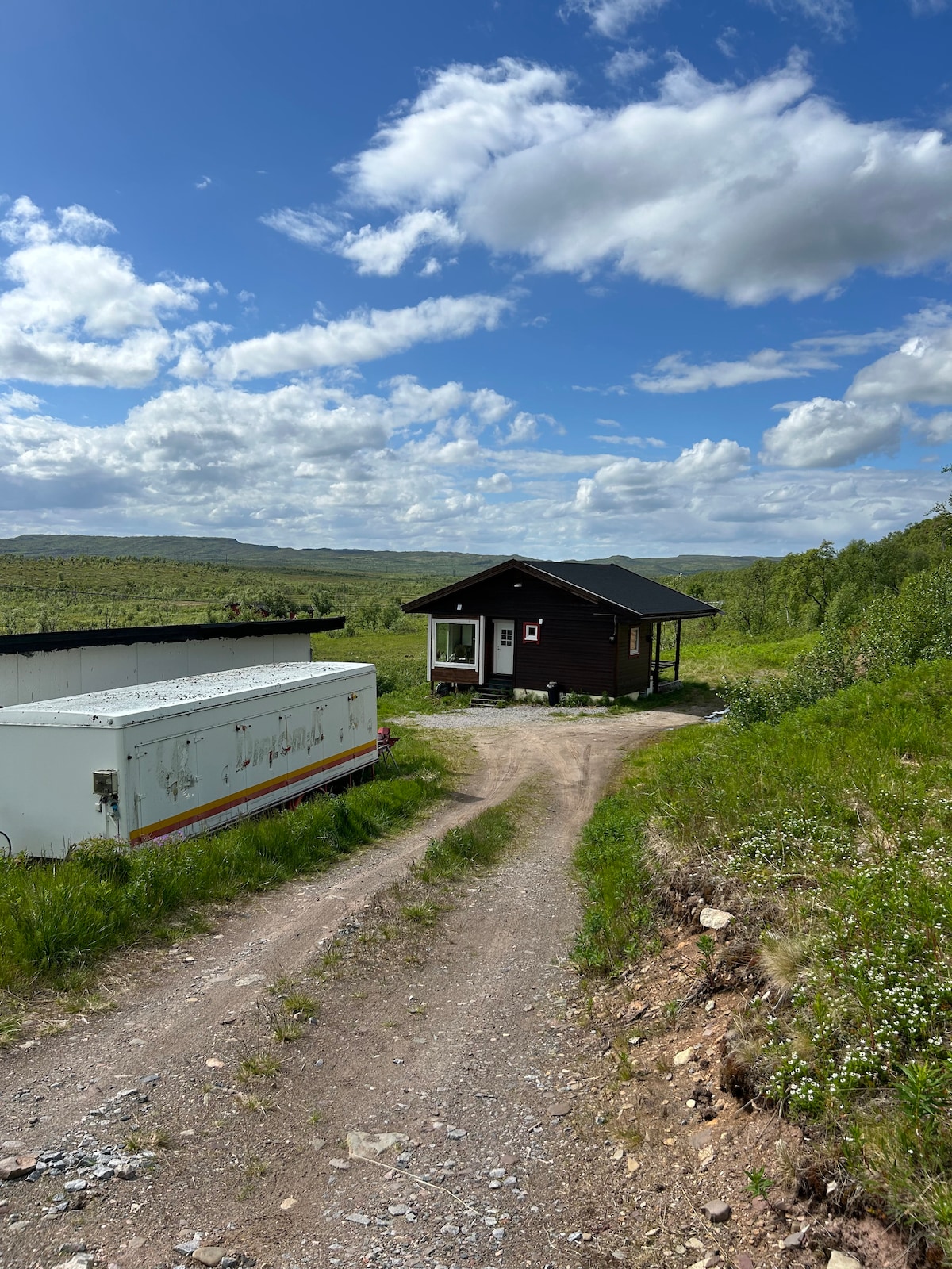 A gravel path leads towards a cozy house set against a backdrop of rolling greenery and blue sky. The exterior of the house features a dark wooden finish and a covered porch, inviting tranquility into the natural landscape.