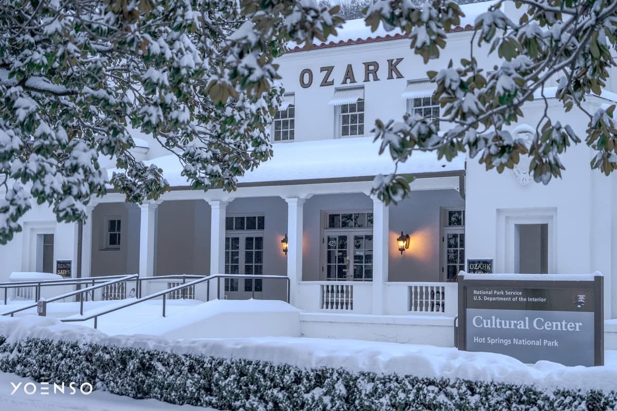 The Cultural Center of Hot Springs National Park is shown in a winter setting, with snow covering the ground and rooftops. The building features a classic design, with white columns and a wide staircase leading to the entrance. A sign indicating the center is prominently displayed.