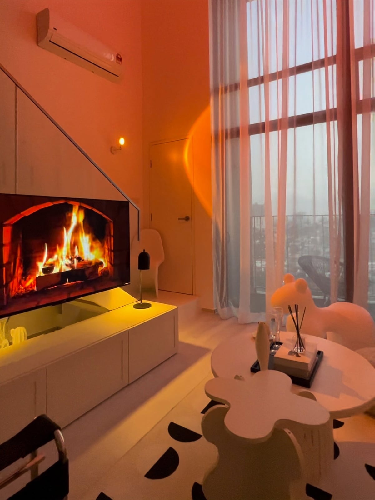 A cozy living area is illustrated with a large television displaying a fireplace. Soft fabric curtains filter natural light, creating a warm ambiance. A modern coffee table and decorative items are positioned on a patterned rug, contributing to an inviting atmosphere.