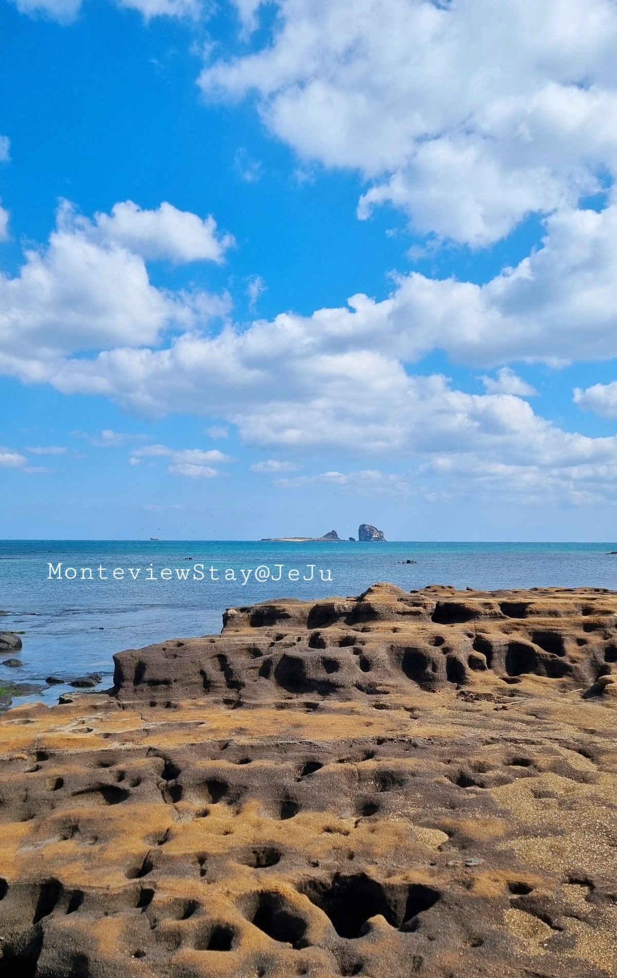 The image displays a scenic view of rocky coastal terrain extending towards the sea, characterized by unique formations and textures. In the distance, an island can be seen against a backdrop of a bright blue sky dotted with white clouds, creating a serene coastal atmosphere.