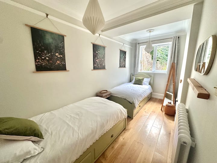 A calming green twin bedroom with two single beds, this room is peaceful and minimal for adult occupancy, while also having features like a small children's bookshelf & toy basket, & removable magnetic wall lights for smaller guests.