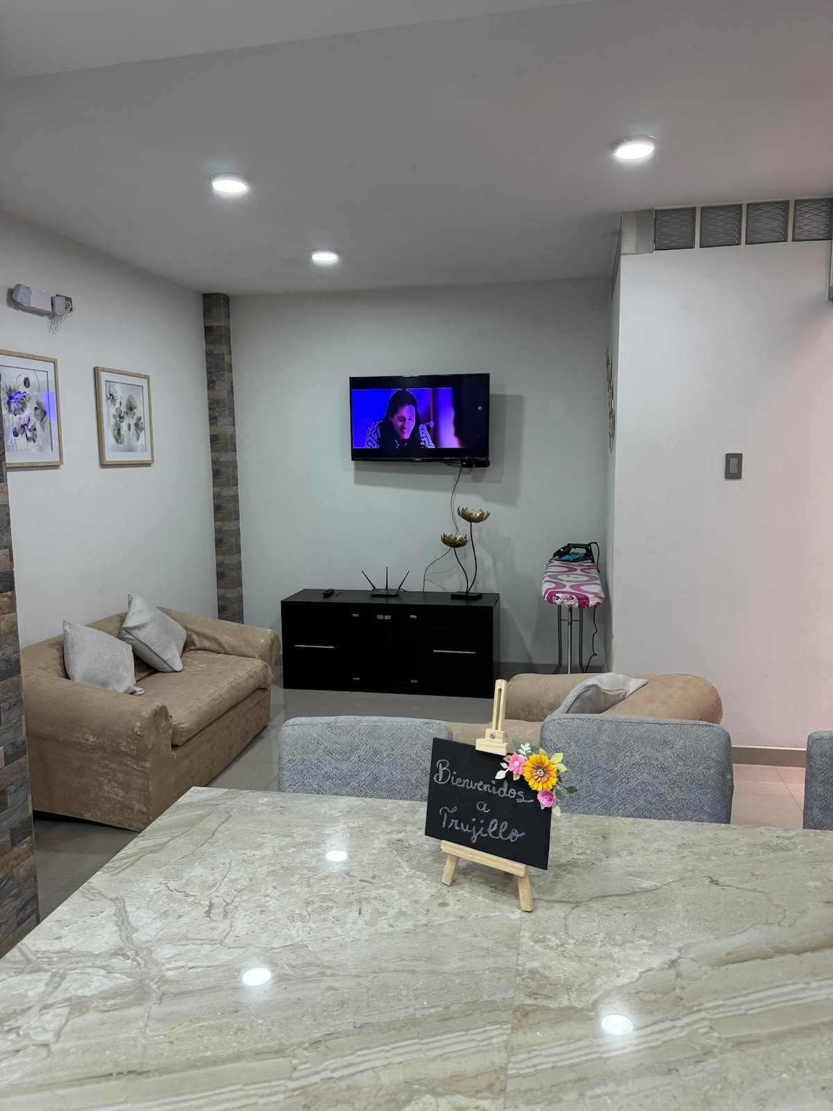 A modern living area is depicted with two comfortable sofas and a sleek television mounted on the wall. Natural light enhances the space, while a decorative sign welcomes guests. A stylish table with a marble top is positioned in the foreground.