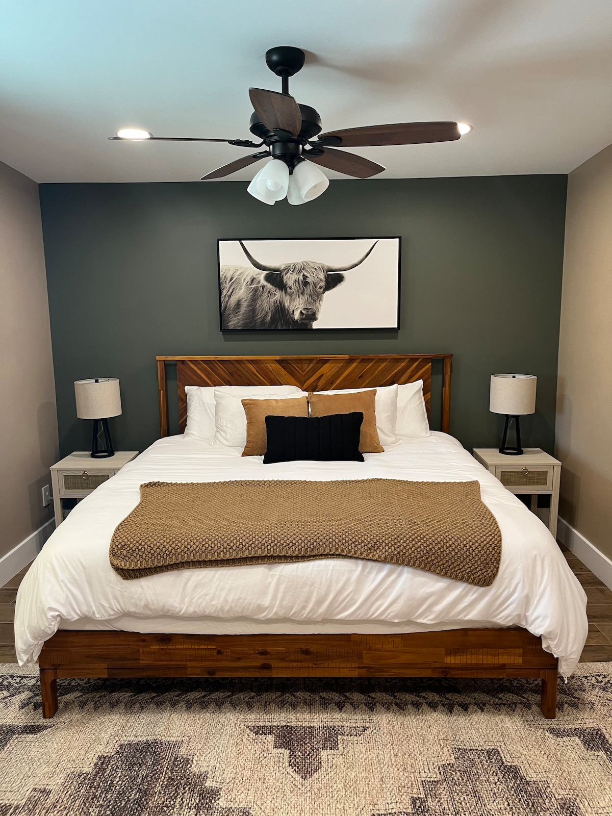 The bedroom features a wooden bed frame with a plush white bedding set, complemented by a decorative brown throw. Two bedside lamps with white shades are positioned on either side of the bed, and a wall art piece of a cow is prominently displayed above.
