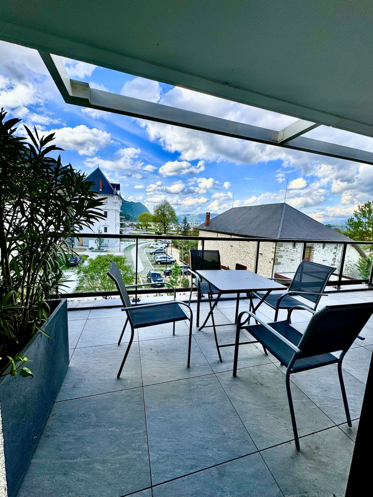 A spacious terrace features a glass railing and is furnished with a table and four chairs. Potted greenery adds a natural touch. The view showcases the surrounding architecture and distant mountains under a partly cloudy sky.