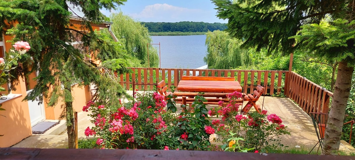 A deck area is framed by vibrant flowers, featuring a wooden table and seating arranged for outdoor gatherings. The serene lake view is visible through lush greenery, creating a peaceful backdrop for relaxation or social activities.