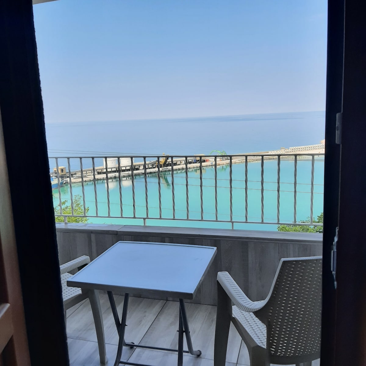 The image captures a serene view from a balcony overlooking a calm harbor. A small table and two chairs are positioned on the balcony, inviting relaxation. The clear turquoise water reflects the sky, with a gentle shoreline visible in the distance.