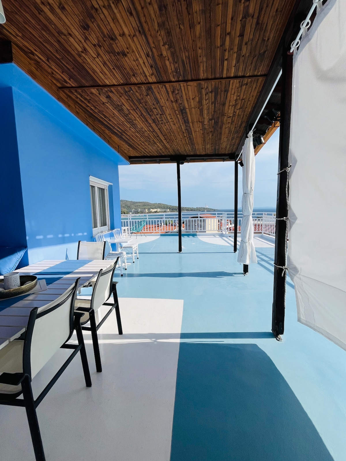 A spacious veranda is featured, offering a wooden ceiling and views of the surrounding landscape. Seating arrangements include several outdoor chairs and tables, set against a backdrop of vibrant blue walls. Light flows across the floor, creating a bright and open atmosphere.