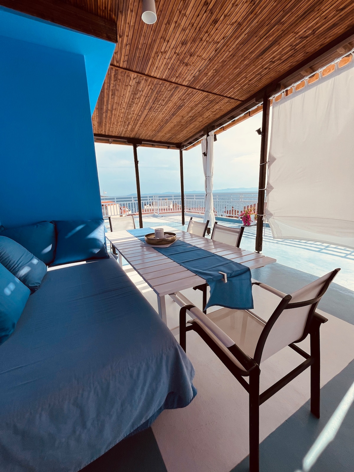 A large veranda is furnished with a dining table surrounded by chairs and a comfortable sofa. Sunlight casts gentle shadows on the blue walls, while sheer curtains flutter in the breeze. The spacious area invites relaxation, offering a view of the surrounding landscape.