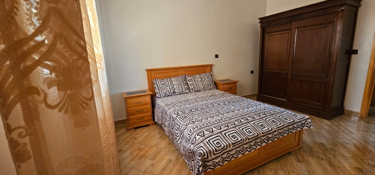 A spacious bedroom is shown, featuring a large bed with a patterned bedspread. Matching nightstands are located on either side of the bed, and a large wooden wardrobe stands against the wall. Soft natural light enters through a window adorned with light fabric curtains.
