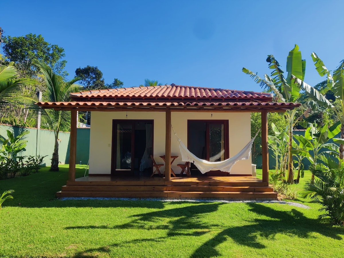 A single-story house is presented with a red-tiled roof, featuring a spacious porch and a hammock. Lush greenery surrounds the property, with palm and banana trees enhancing the outdoor area. Large windows allow natural light to fill the interior.
