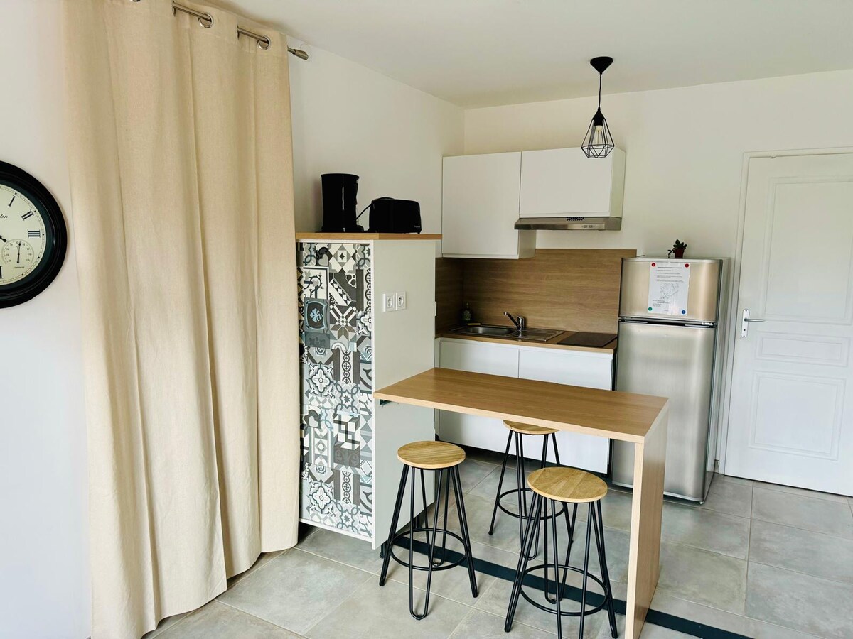 A modern kitchen area features a light wood dining table with two stools. White cabinets and a stainless steel refrigerator are visible, alongside a small sink and cooking area. A decorative curtain softly separates the space, while a large clock adorns the wall.
