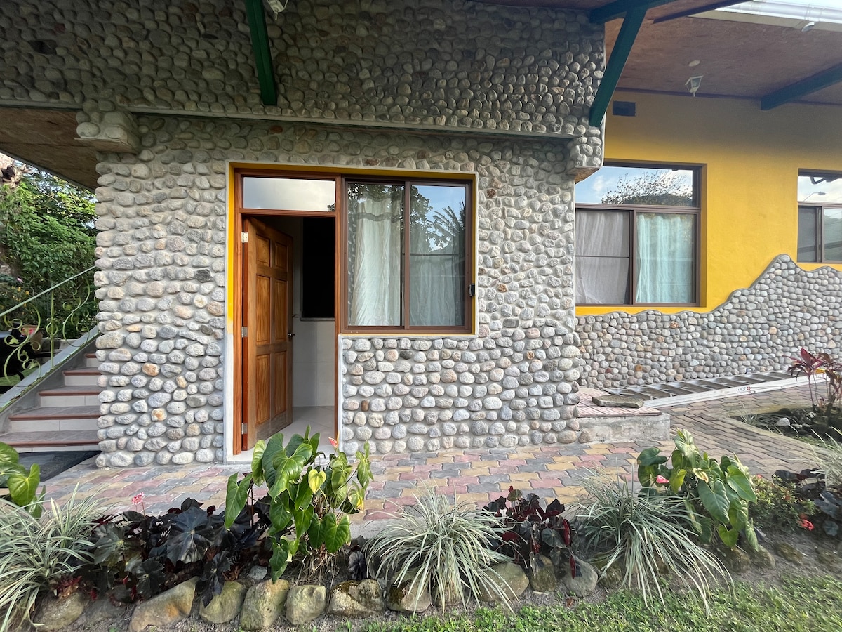 The entrance to the private suite is framed by a stone exterior, showcasing a blend of natural textures. A large window allows natural light to enter, while colorful landscaping featuring various plants enhances the welcoming atmosphere of the space.