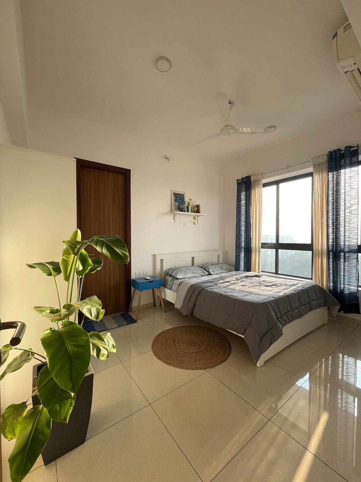 A bright bedroom features a spacious white bed with a gray blanket laid across it. A potted plant adds a touch of greenery beside the bed. Tall windows with sheer curtains allow natural light to fill the room, and a woven rug sits on the tiled floor.