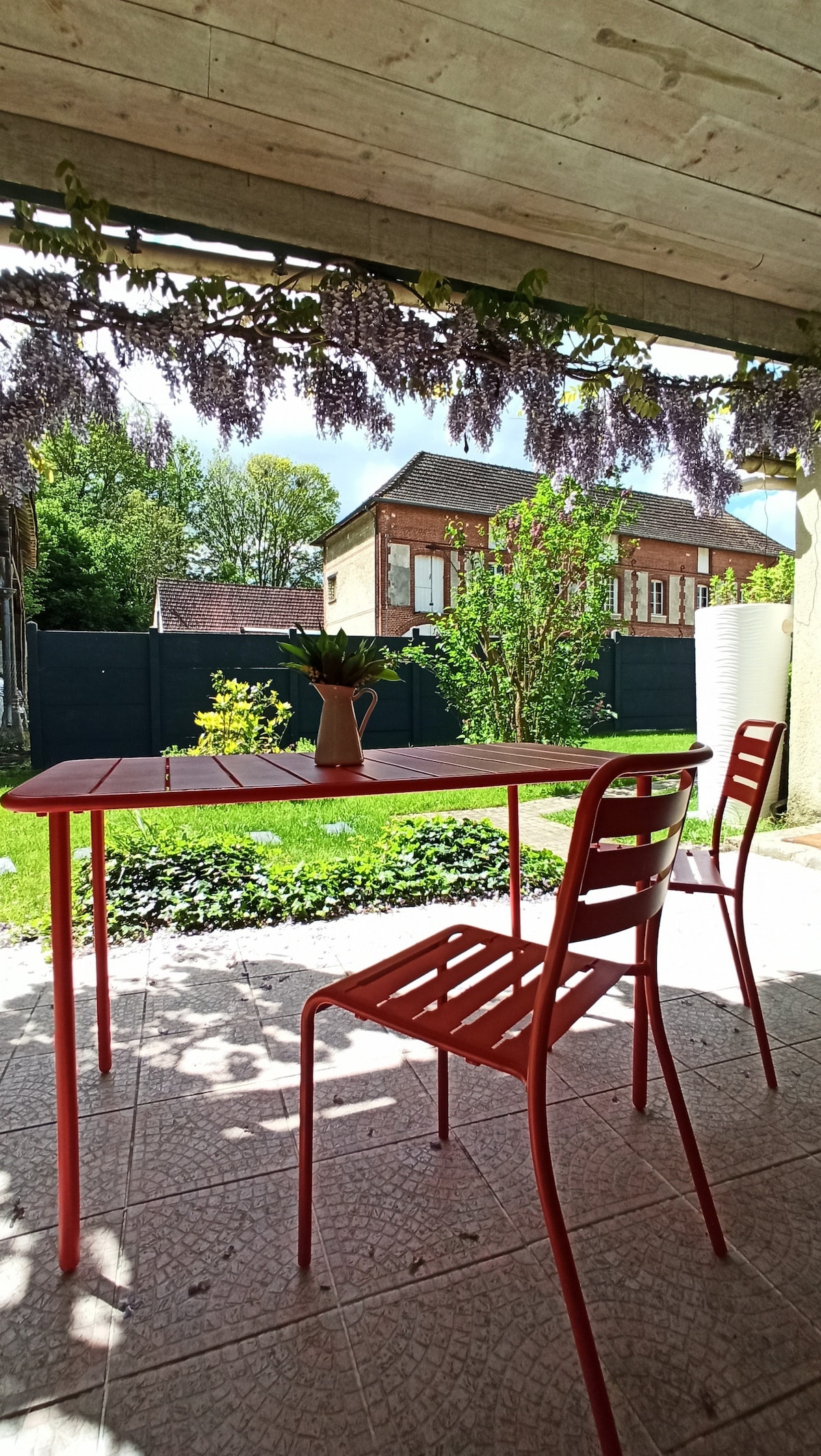 An outdoor patio area is featured, with a bright red table and two matching chairs. Overhead, lush wisteria blooms create a charming canopy. The garden beyond is visible, characterized by greenery and flowering plants, providing a tranquil outdoor setting.