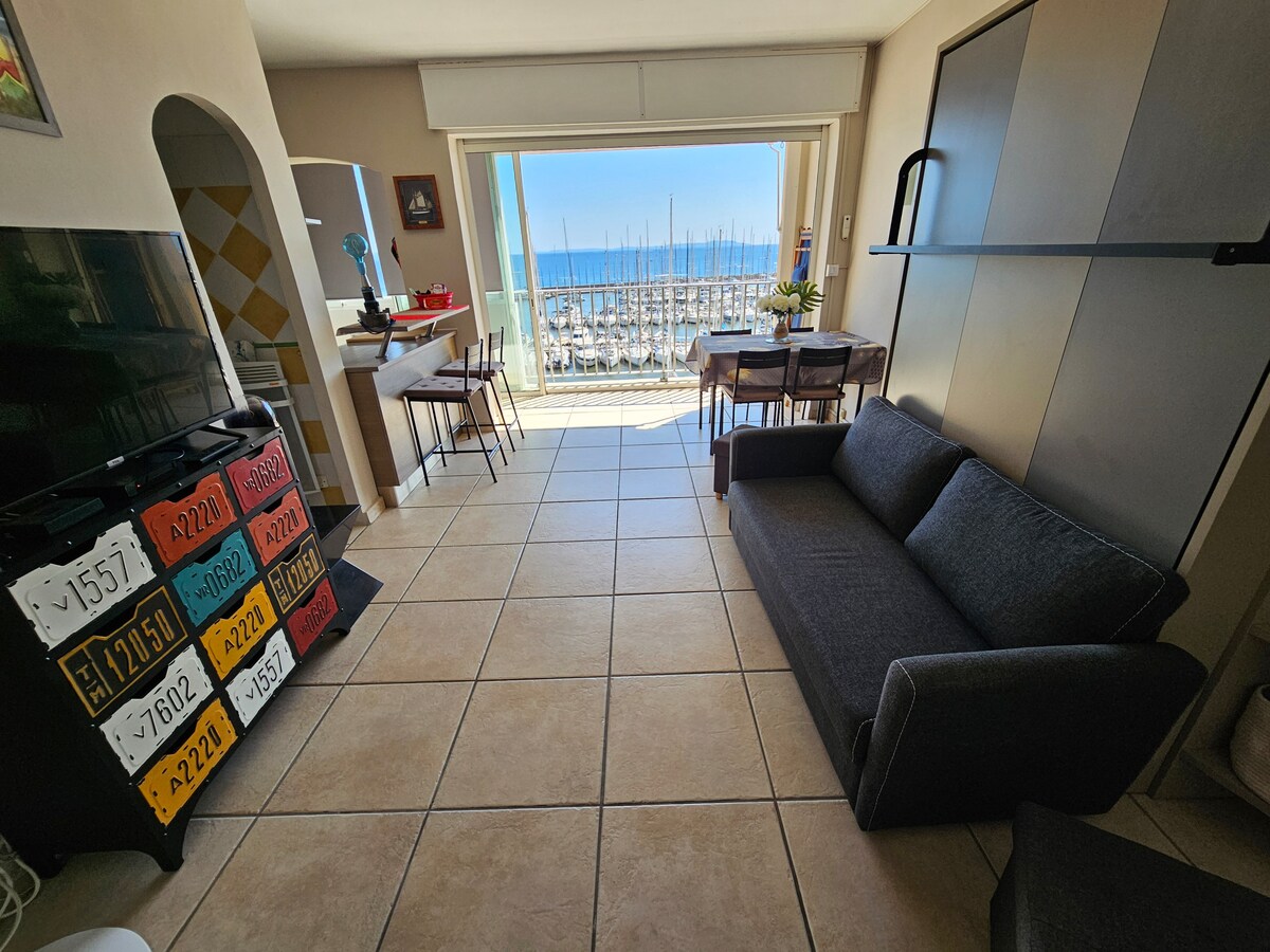 The living area features a comfortable sofa oriented toward large glass doors, which reveal a stunning view of the port and the ocean. A dining table with chairs is positioned nearby, and a television is mounted on the wall, completing the open layout.