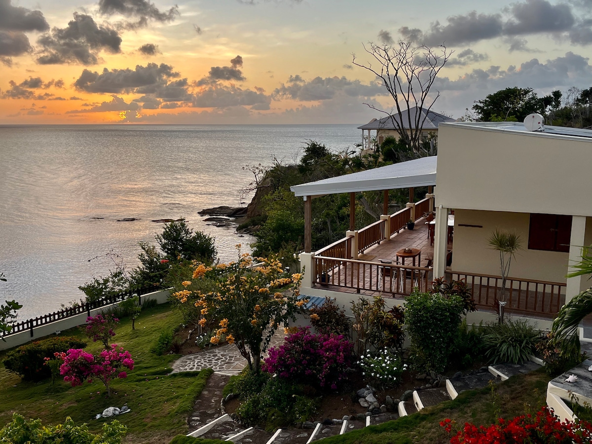 A stunning sunset is captured over the calm ocean, illuminating the sky with soft hues. The guest house features a spacious veranda with seating, surrounded by lush gardens filled with colorful flowers and tropical plants. The scenic view showcases the tranquil coastal landscape.