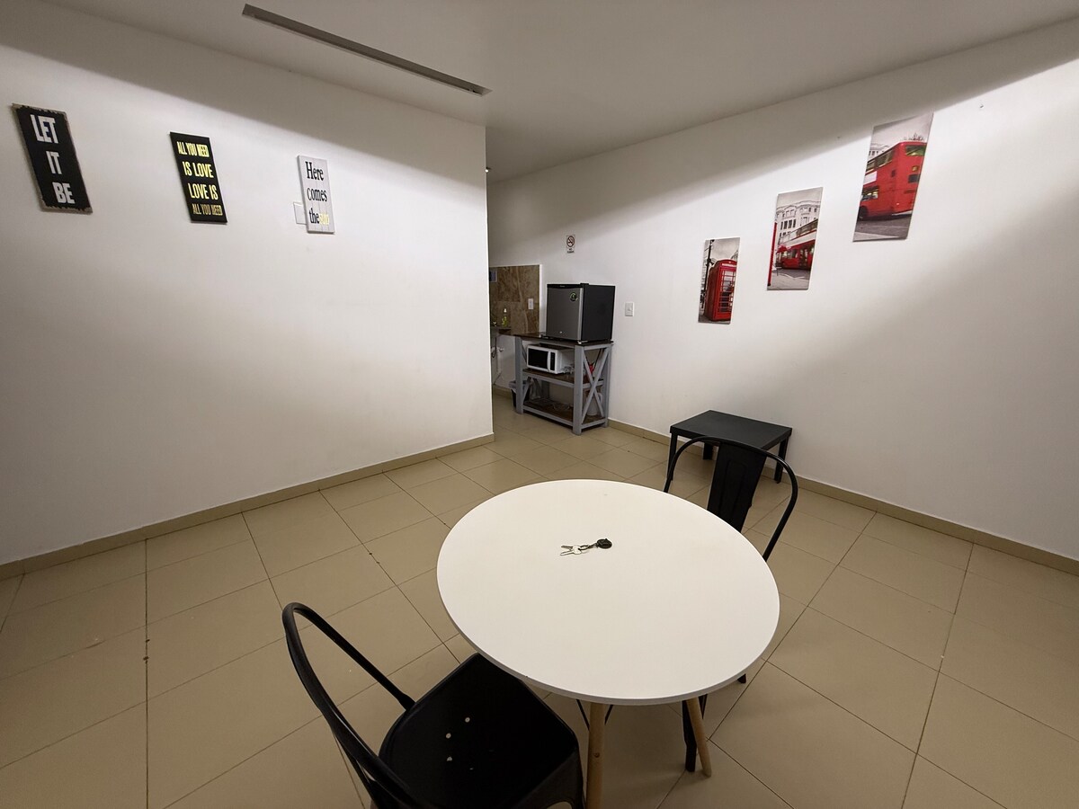 An open area is furnished with a round white table at the center and black chairs around it. Light-colored walls are adorned with framed artwork, and a kitchen area is visible in the background, featuring a wooden cabinet and a countertop.