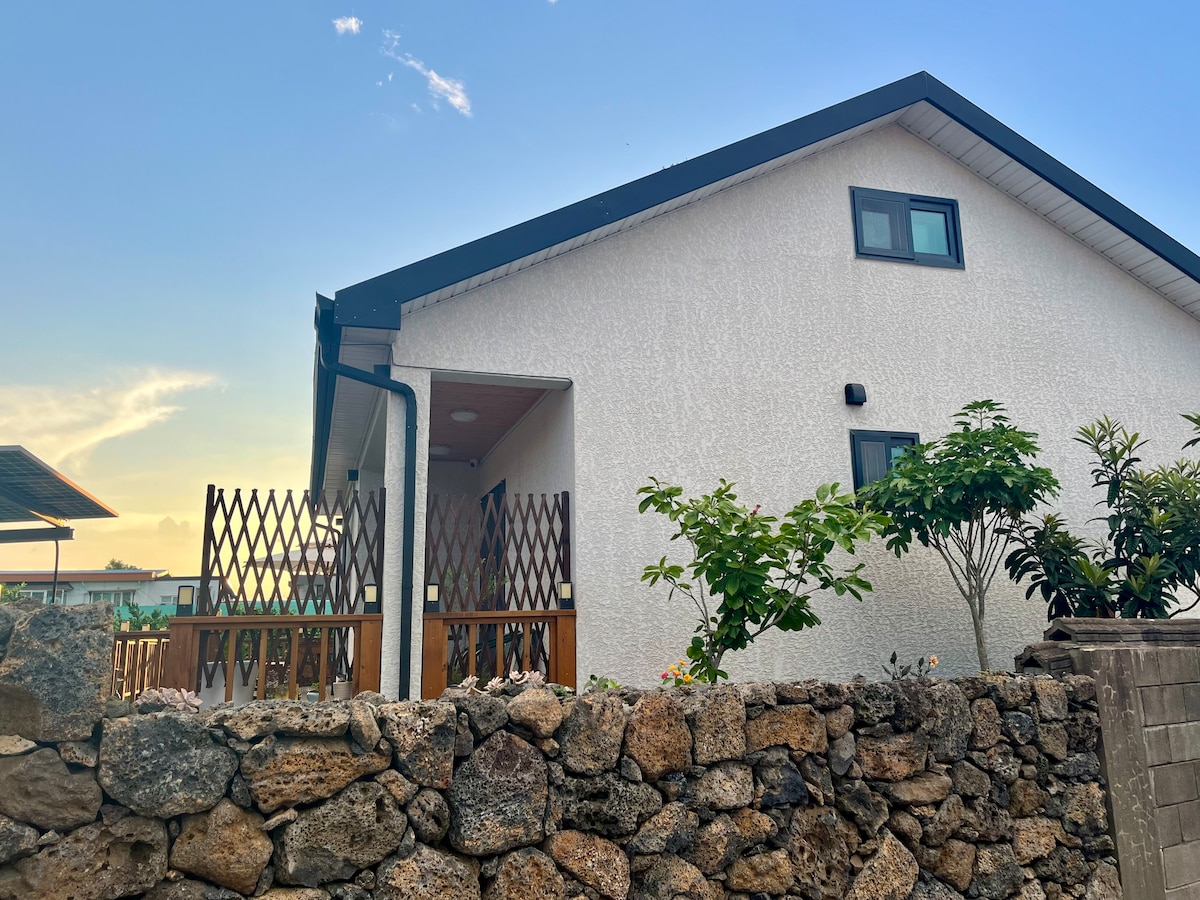 The exterior of a single-story house is depicted, featuring a clean white wall and a gabled roof. A wooden deck adorned with plants sits at the entrance. A low stone wall surrounds the property, contributing to a rustic charm against a backdrop of a clear sky.