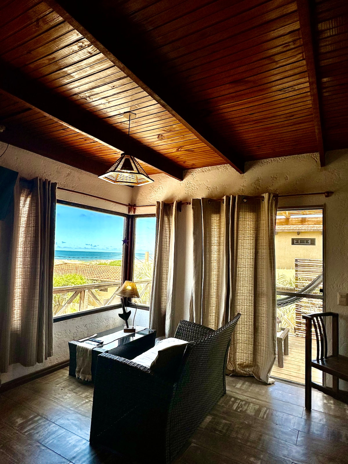 A comfortable seating area features a woven chair and side table, with large windows showcasing expansive ocean views. Natural light filters through sheer curtains, illuminating the wooden ceiling that enhances the cozy atmosphere.