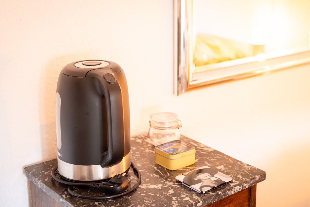A kettle and a small jar sit on a marbled nightstand, accompanied by a decorative tin and a photograph. A mirror with a simple frame reflects soft light, enhancing the serene environment.