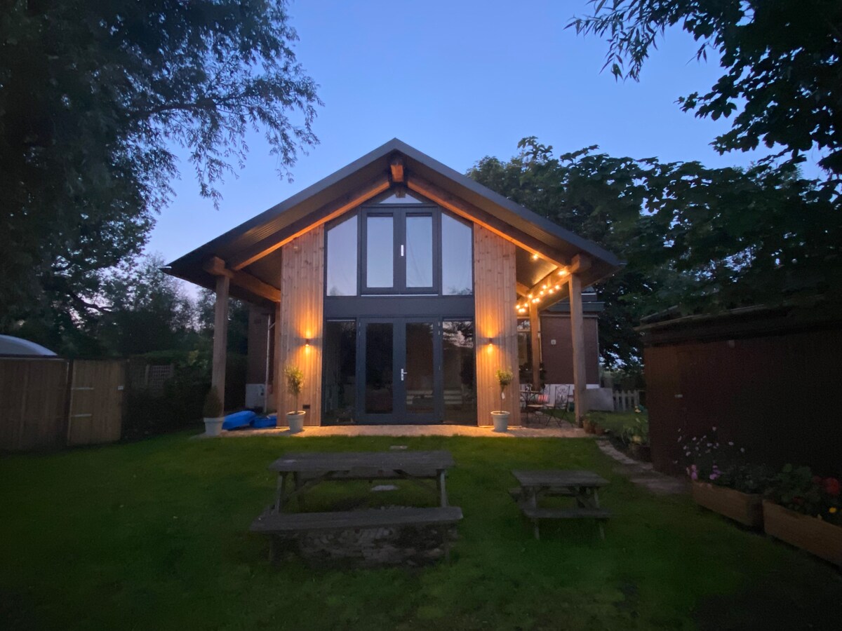 A wooden house with large glass doors is set in a lush garden, softly illuminated by outdoor lights. The structure features a sloping roof and is surrounded by trees, creating a serene and inviting atmosphere. Outdoor seating areas are available on the lawn.