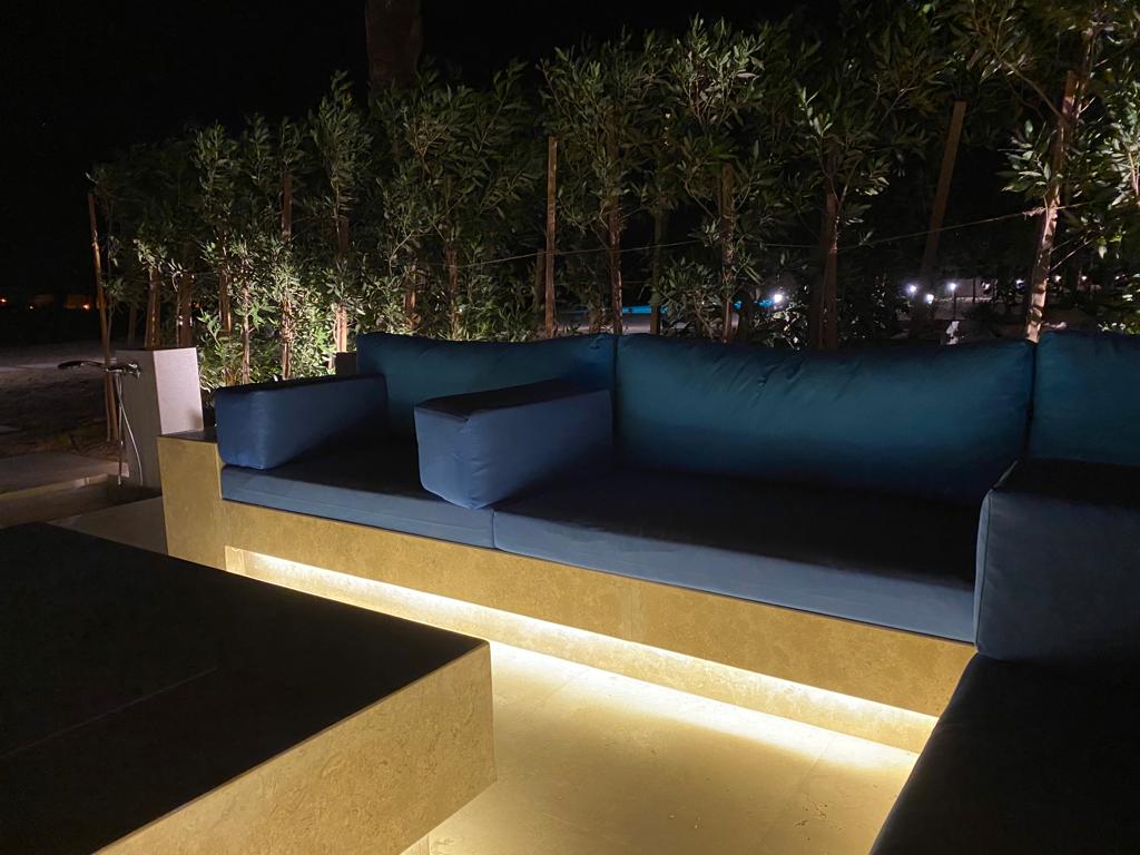 A modern seating area features a deep blue couch situated against a backdrop of lush greenery. Soft lighting illuminates the space from below, creating a cozy ambiance. The environment suggests an inviting setting for relaxation during evening hours.