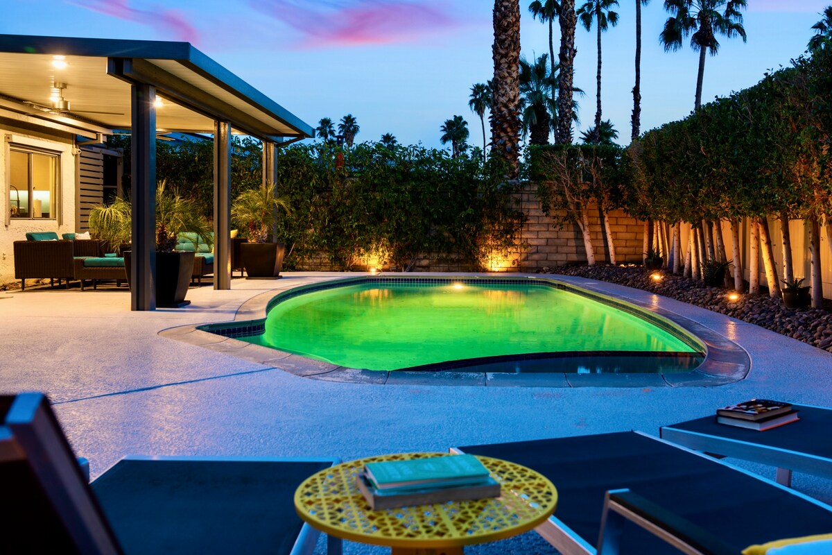 A serene backyard features a glowing green pool surrounded by smooth concrete. Comfortable lounge chairs with side tables are positioned nearby. Lush greenery and palm trees provide privacy, while soft lighting accentuates the space during twilight.