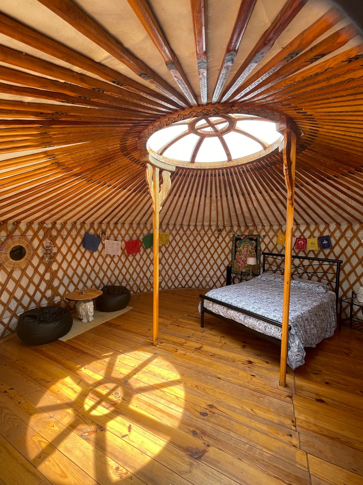 An inviting yurt interior showcases a spacious bed with a patterned cover. Natural light filters through a circular skylight, creating a soft glow on the wooden floor. Decorative elements include various artworks and a seating area with two bean bags and a low wooden table.