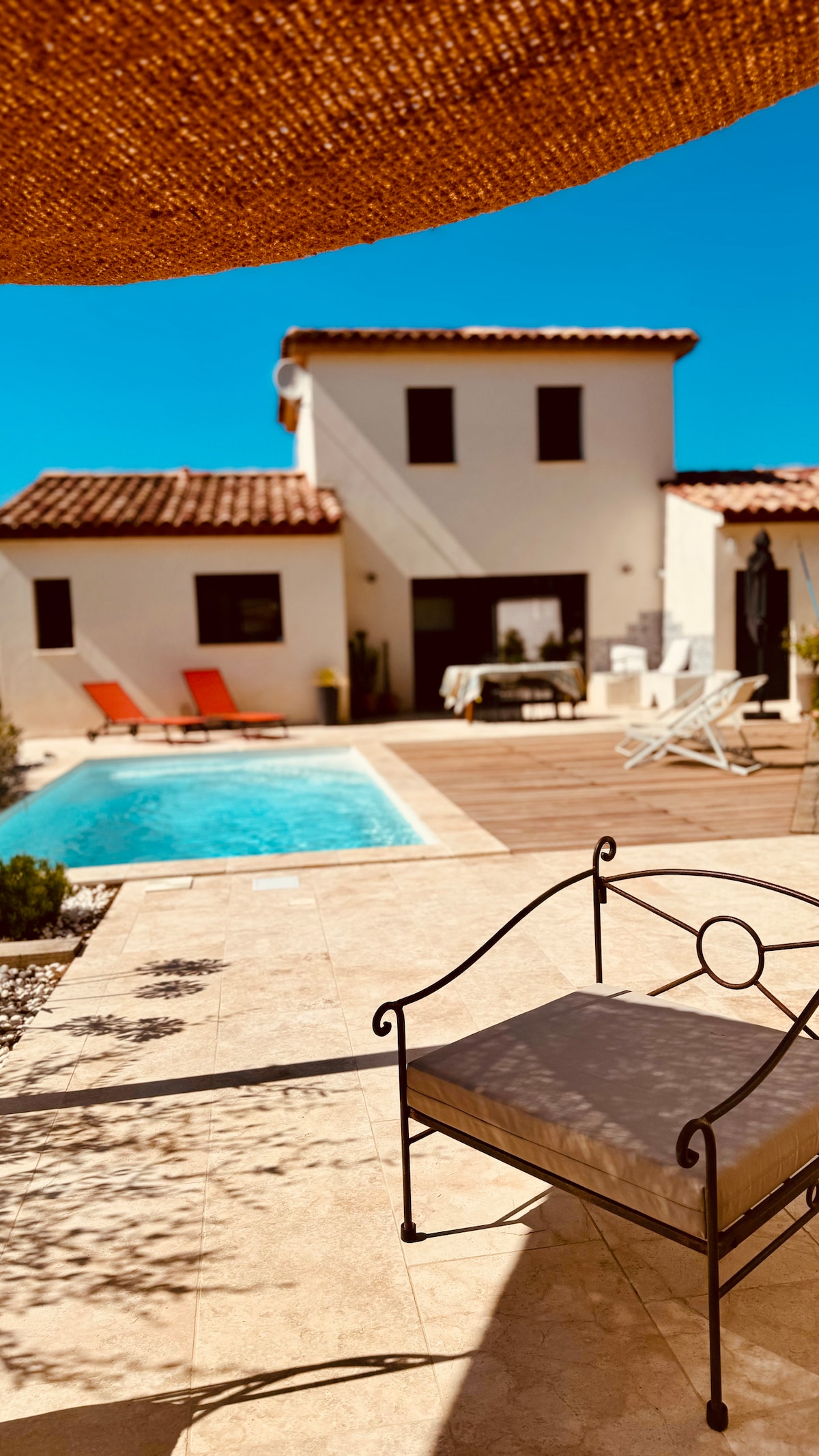 A view of a modern villa featuring a well-maintained pool surrounded by a wooden deck. Sun loungers and orange chairs are positioned near the water, while a shaded seating area with a metal frame chair is visible in the foreground.