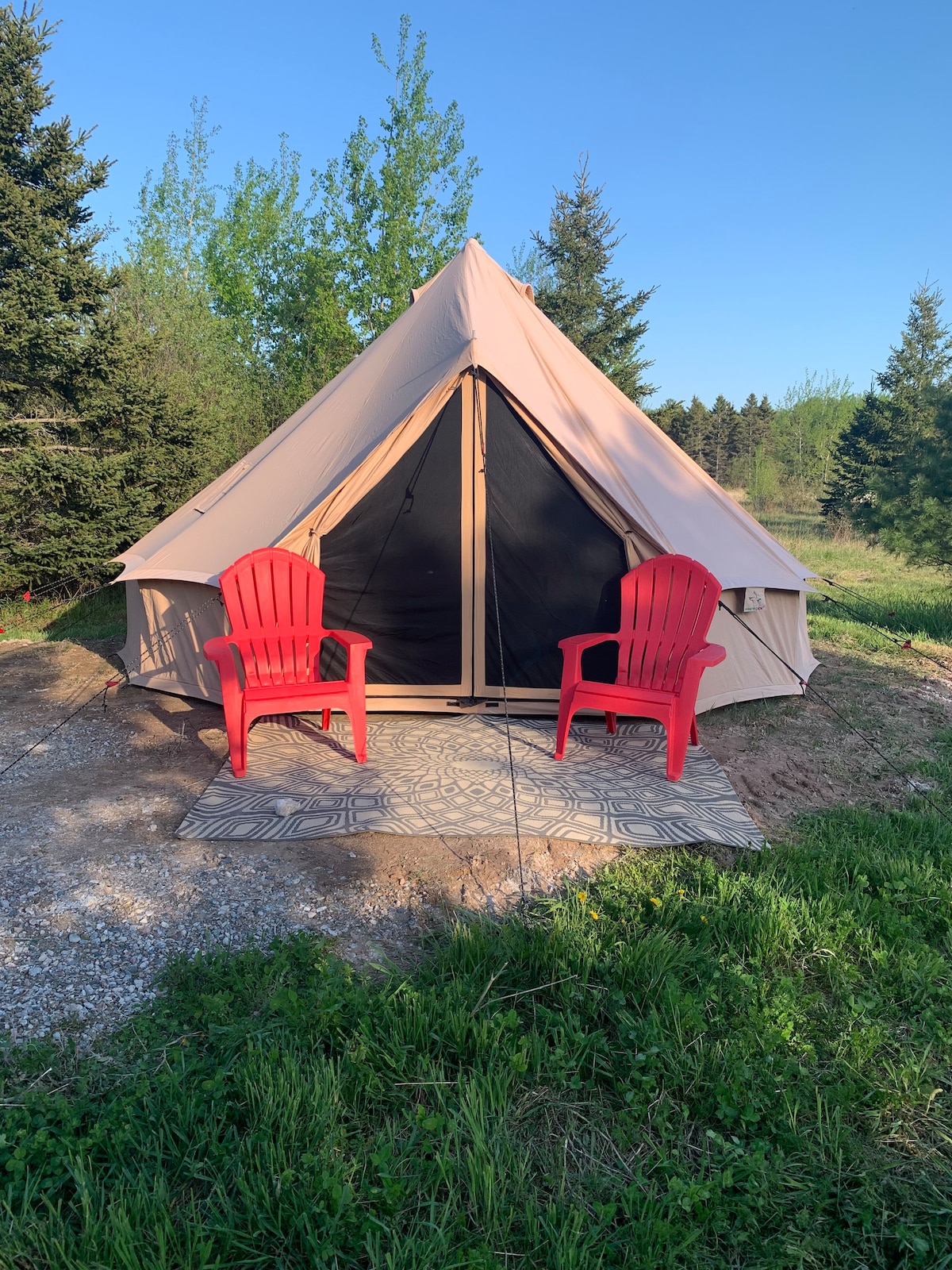 A spacious glamping tent is set against a natural backdrop, featuring a screened entrance and a simple outdoor mat. Two red Adirondack chairs are positioned on the mat, inviting relaxation while enjoying the surrounding greenery.