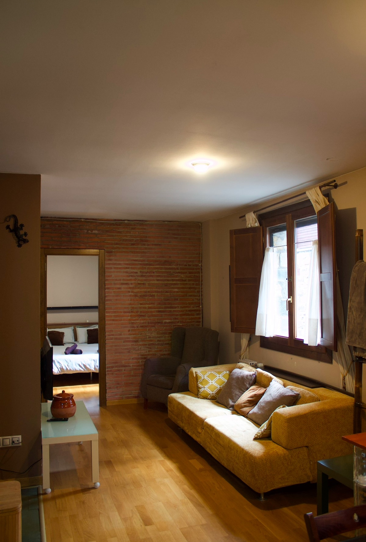 A cozy living space features a yellow sofa adorned with various cushions, positioned near a glass coffee table. Large windows allow natural light to fill the room, highlighting the warm wood flooring. A dark armchair and an inviting mirror are also visible in the area.