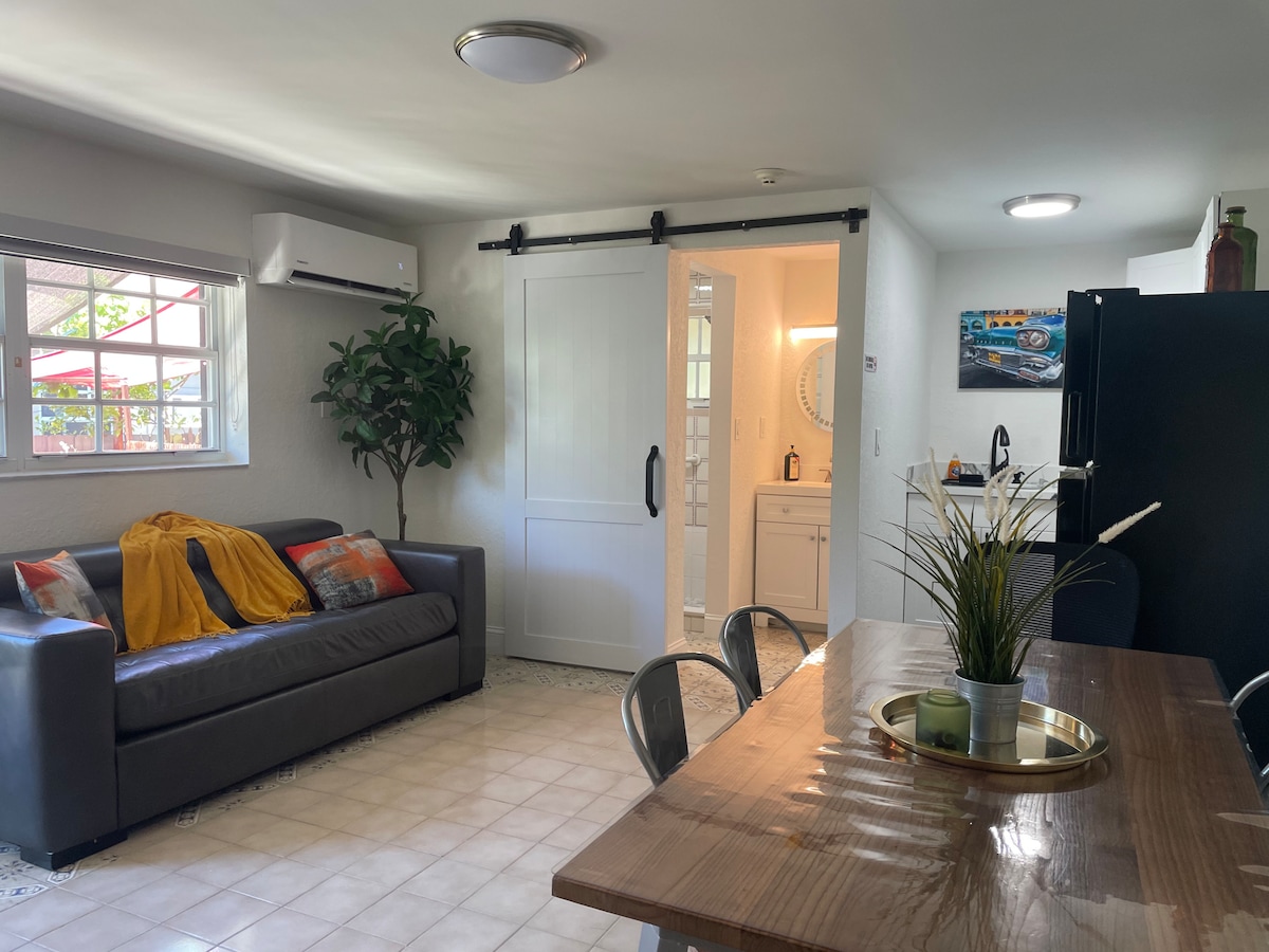 A comfortable living area features a dark sofa adorned with colorful cushions, a yellow throw draped over one arm, and a potted plant nearby. A dining table with metallic chairs is positioned close to the kitchen, which includes a black refrigerator, and a sliding door offers access to the bathroom.