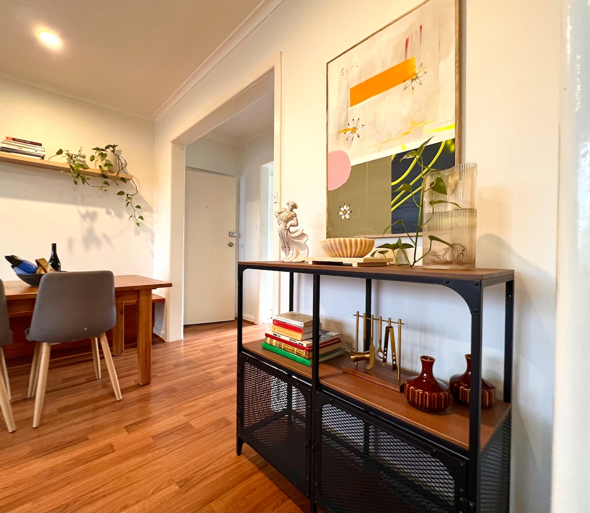 A stylish living space is displayed, featuring a contemporary shelving unit adorned with decorative items and books. A dining table with sleek chairs is positioned nearby, and a colorful wall art piece adds visual interest. Natural light enhances the warm wood flooring.