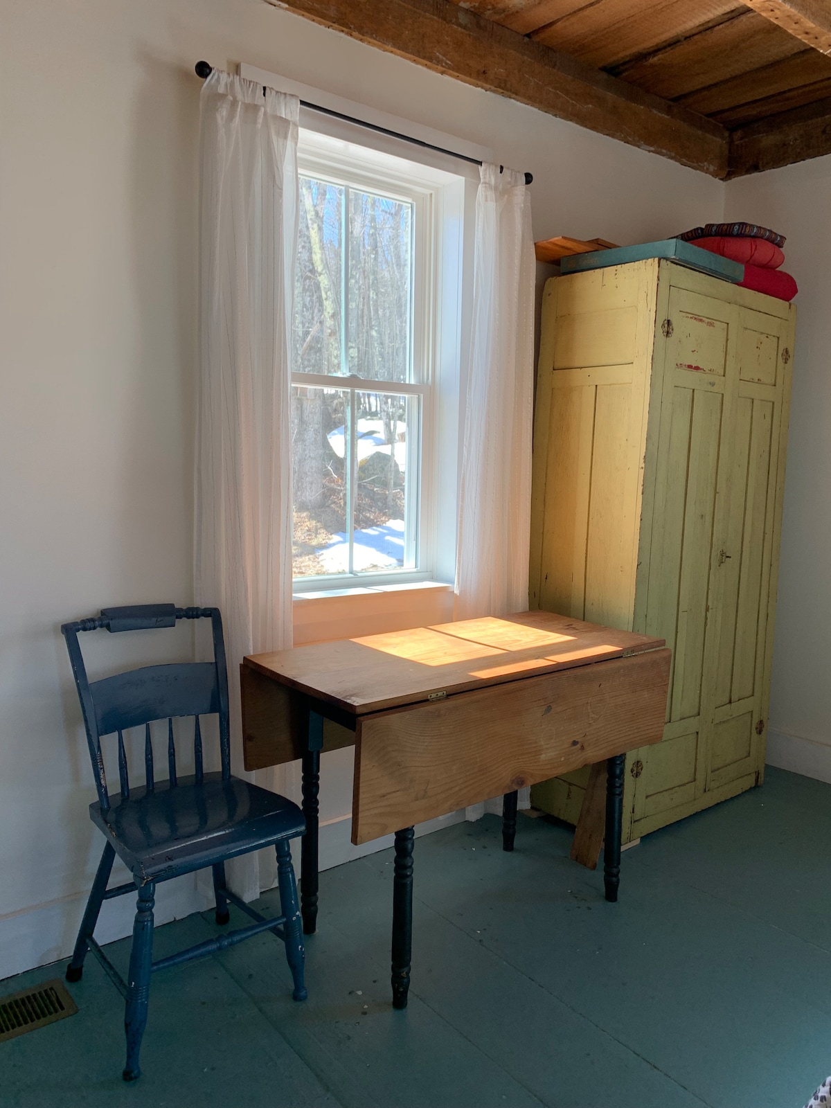 A small workspace is present, featuring a simple wooden table with sunlight illuminating its surface. A blue chair is positioned next to the table. An antique yellow cupboard stands nearby, providing additional storage. A window with light curtains allows natural light to fill the space.