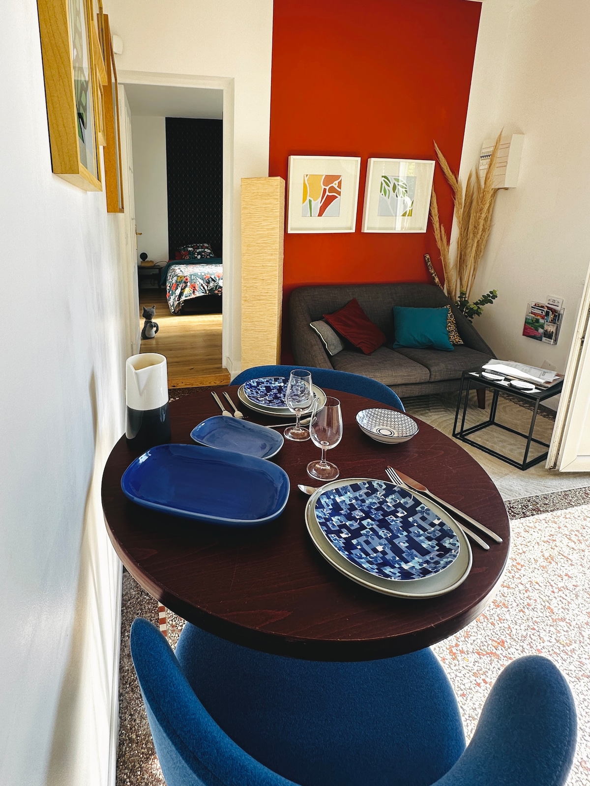 A cozy dining area features a round table set with blue and white plates, accompanied by elegant glassware. A comfortable blue chair sits beside the table, with a sofa and wall art in the background, creating a welcoming atmosphere.