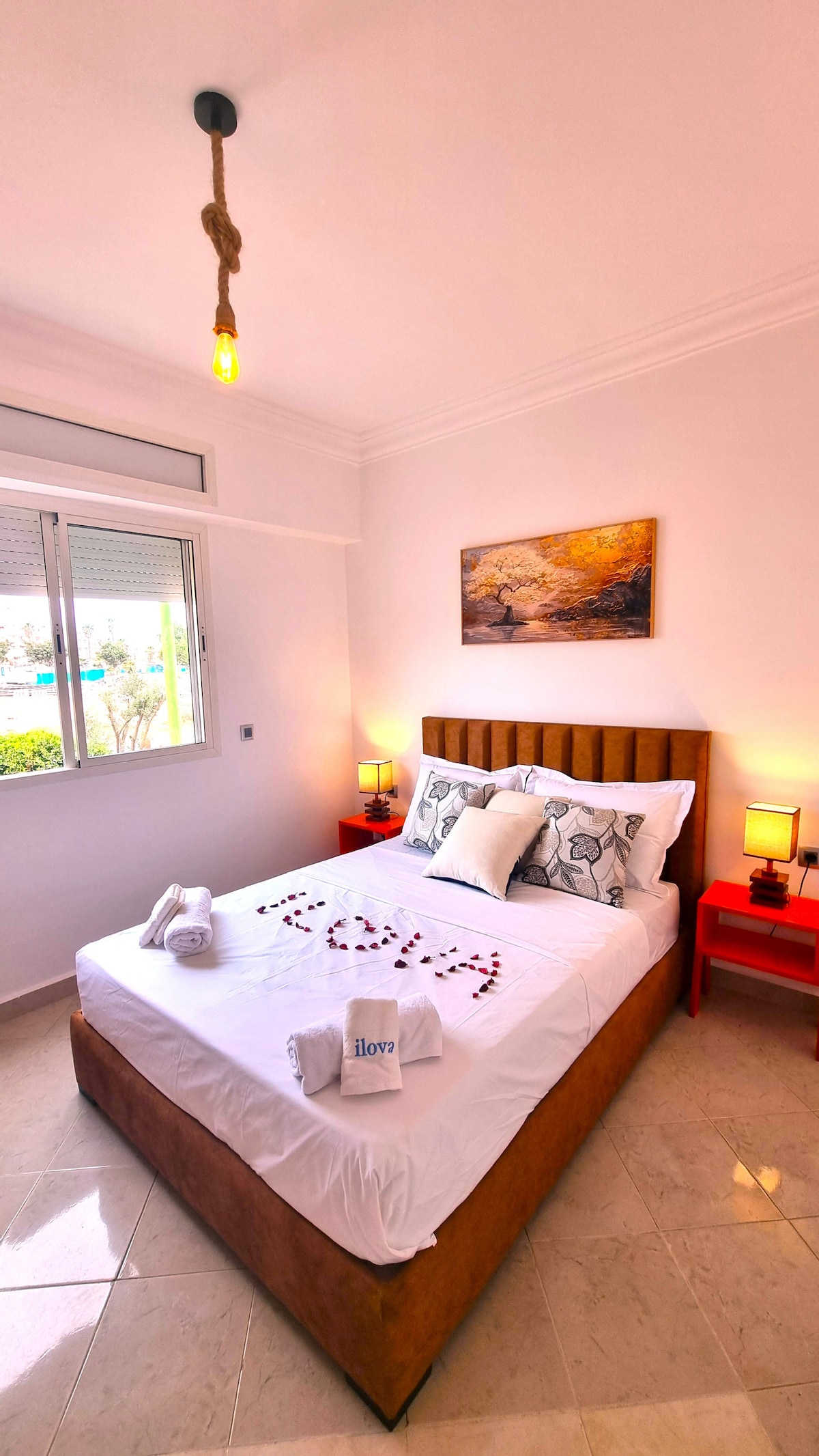 הירגעו בחדר השינה הבהיר והמזמין הזה, עם מצעים נקיים, תאורה רכה, שידה ליד הנוחה ואור שמש טבעי שזורם דרך החלון עם נוף יפה.