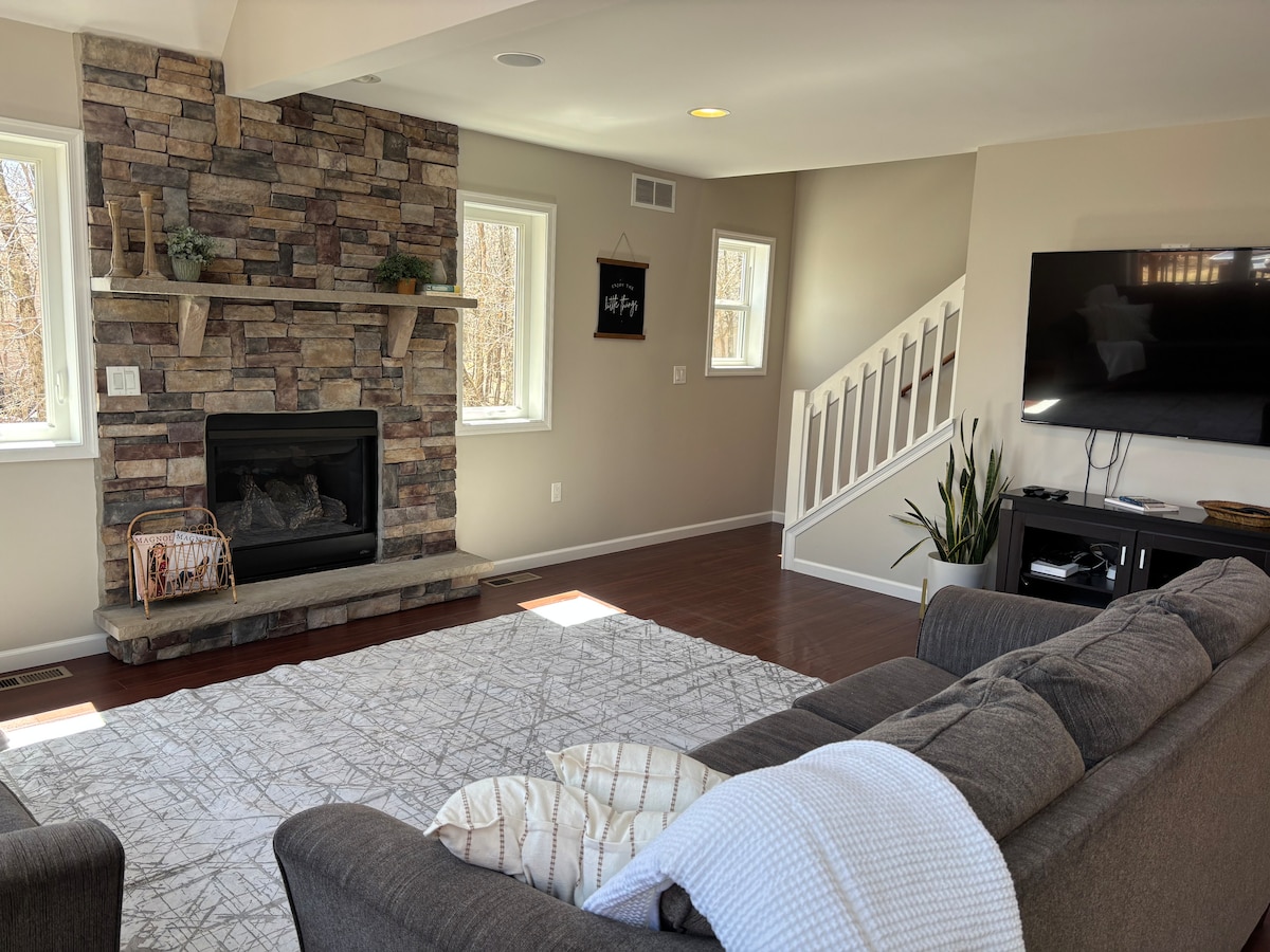 A spacious living area features a stone fireplace as its focal point, surrounded by large windows allowing natural light to fill the room. A comfortable gray sofa is positioned on a textured area rug, and a mounted television is visible on a media console.