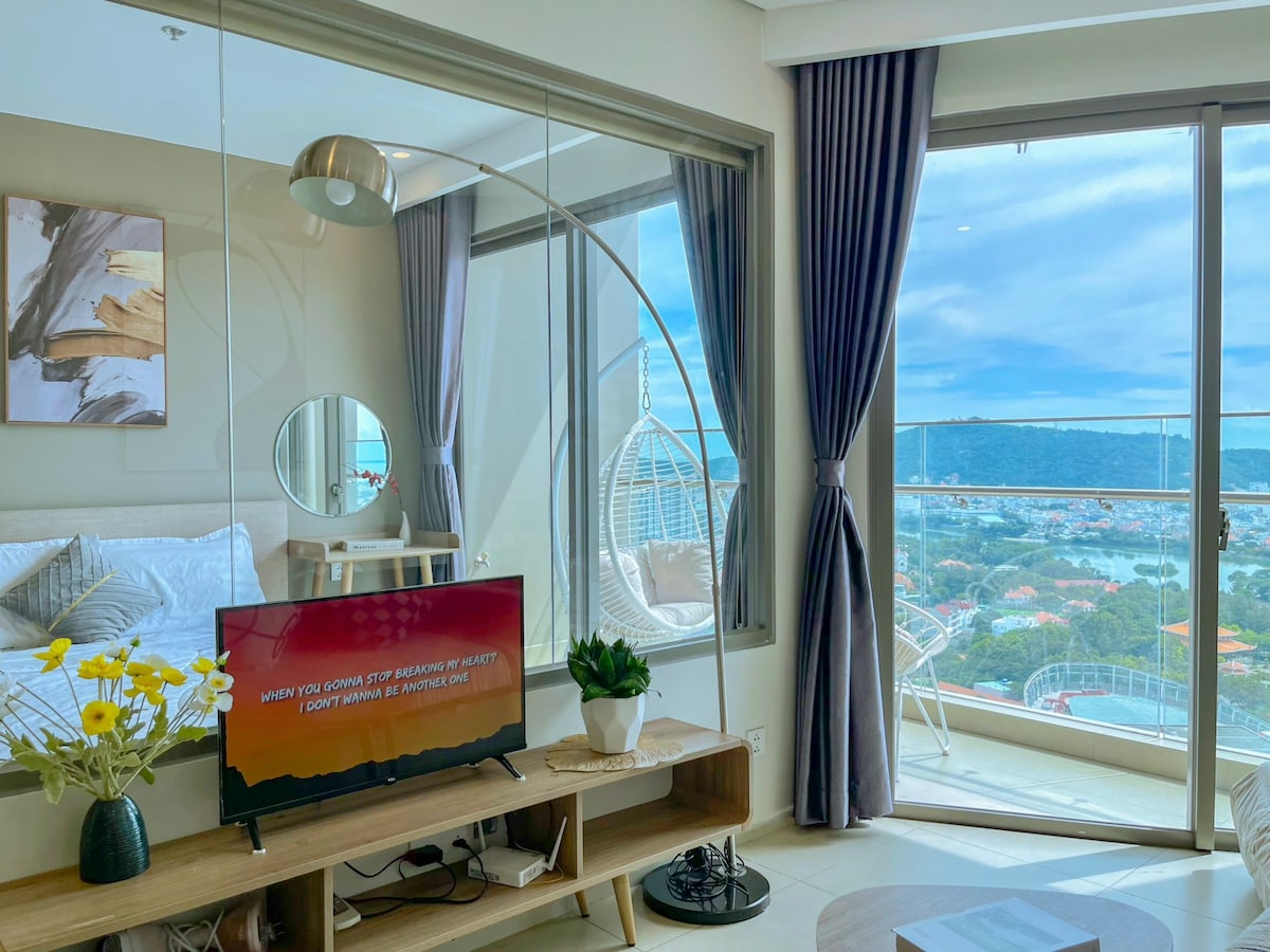 A contemporary living area features a spacious bed framed by soft linens, with a hanging chair beside a large window offering expansive views. A television rests on a wooden stand, complemented by decorative flowers on a side table. Natural light brightens the inviting interior.