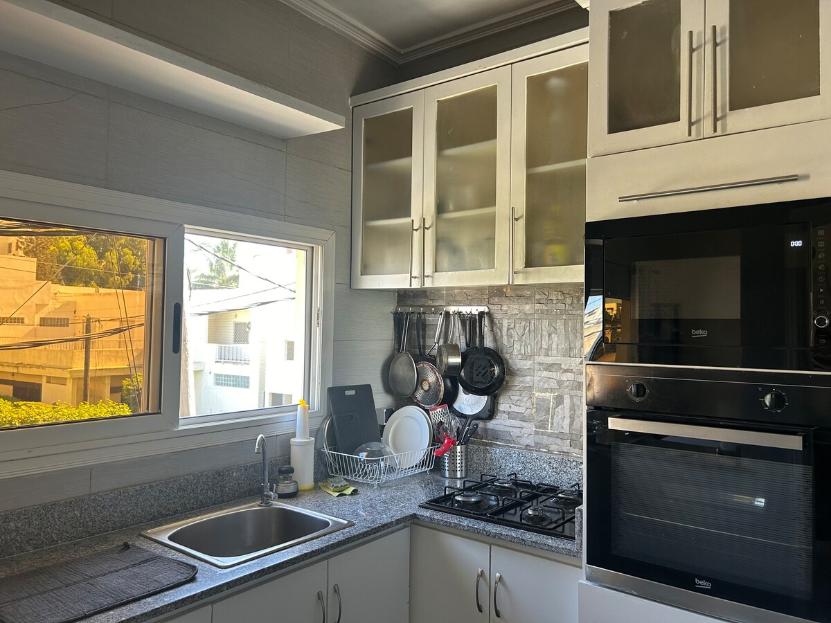 A functional kitchen space is presented, featuring modern appliances and a blend of cabinetry. Natural light filters through a window, highlighting the organized shelves and countertop. Essential utensils and cookware are arranged for convenience, alongside a double sink and a gas stove.