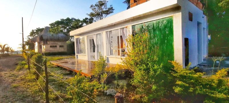 A modern white building is shown, featuring large glass doors that provide natural light. Lush green foliage frames the structure, enhancing its connection with the outdoor environment. A wooden deck extends from the building, allowing for outdoor relaxation.