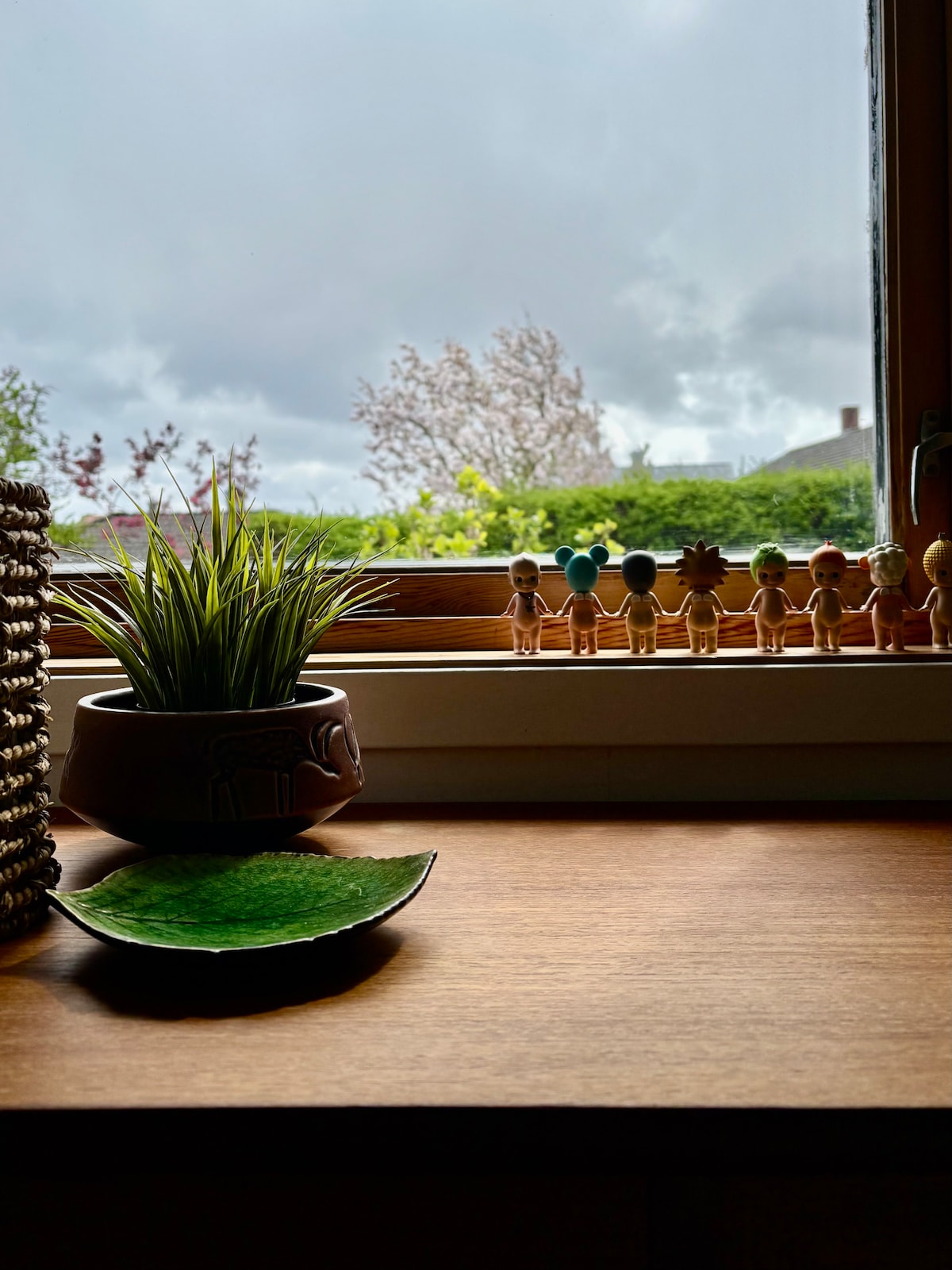 A view is captured from a window, featuring a collection of wooden figures lined on the sill. A small potted plant adds a splash of greenery beside a decorative leaf-shaped plate. The outside scene is partially obscured by clouds and foliage.