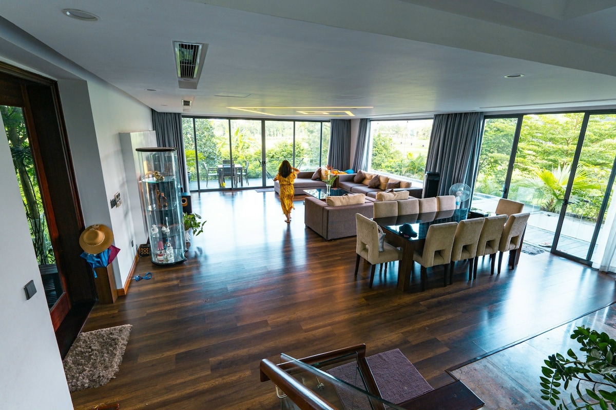 A spacious open-plan living area is depicted, featuring a large sectional sofa and a dining table. Floor-to-ceiling windows provide natural light and views of the surrounding greenery. A glass display cabinet adds an elegant touch, enhancing the modern design of the space.