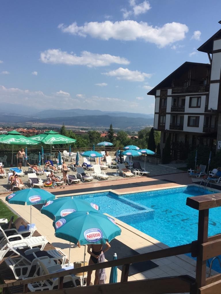 A vibrant outdoor pool area is depicted, surrounded by lounge chairs and umbrellas. The pool reflects the blue sky, with mountains visible in the background. The multi-level building is positioned nearby, complementing the serene setting.