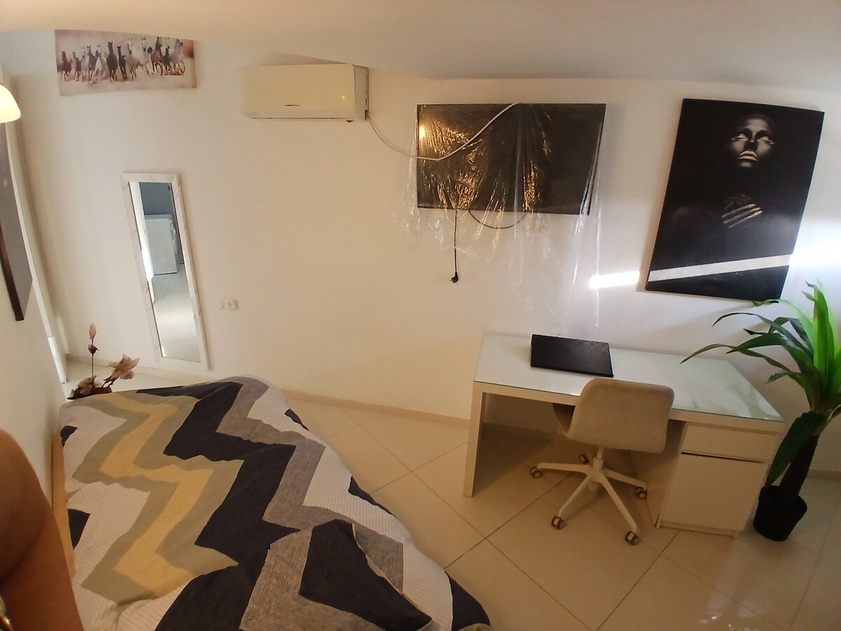 A bright and airy bedroom features a spacious bed dressed in a patterned quilt. A modern desk and chair are positioned next to a large mirror, while a plant adds a touch of greenery. A mounted television is visible on the wall, providing functionality.