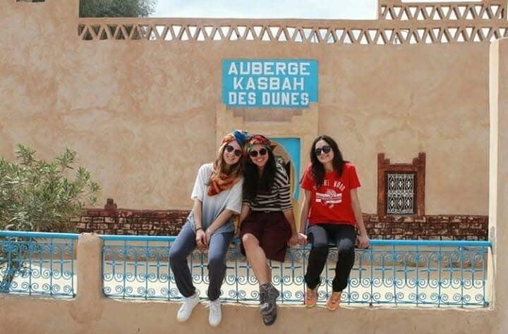A group of three individuals is seated on a decorative blue railing, in front of a building adorned with a sign reading 'Auberge Kasbah des Dunes.' The walls exhibit an earthy tone, complemented by subtle architectural details and a few outdoor plants nearby.