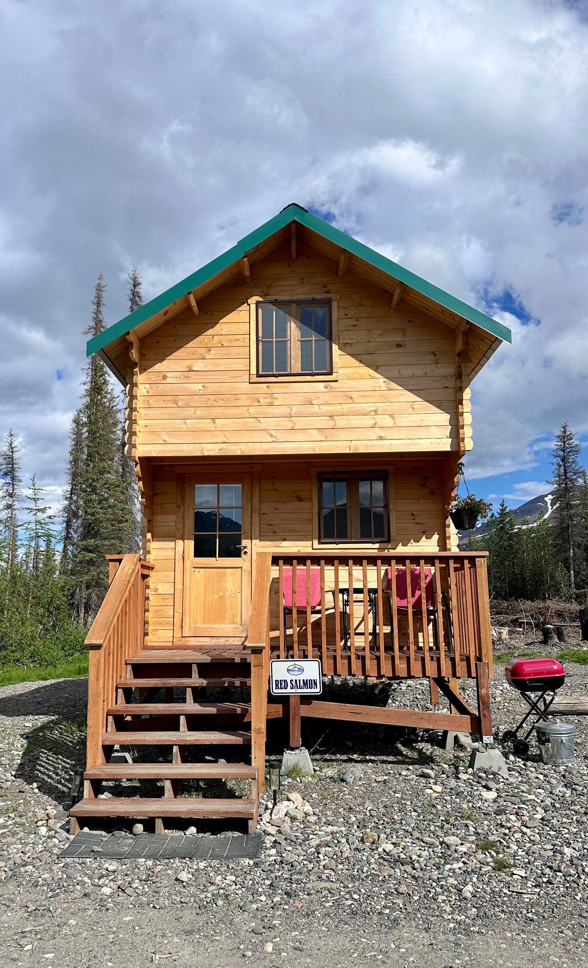 A newly built cabin features a wooden exterior with a sloping roof and turquoise accents. A set of stairs leads to the front door, flanked by windows. The entrance is adorned with a welcoming deck, which includes seating and a small grill, surrounded by natural scenery.