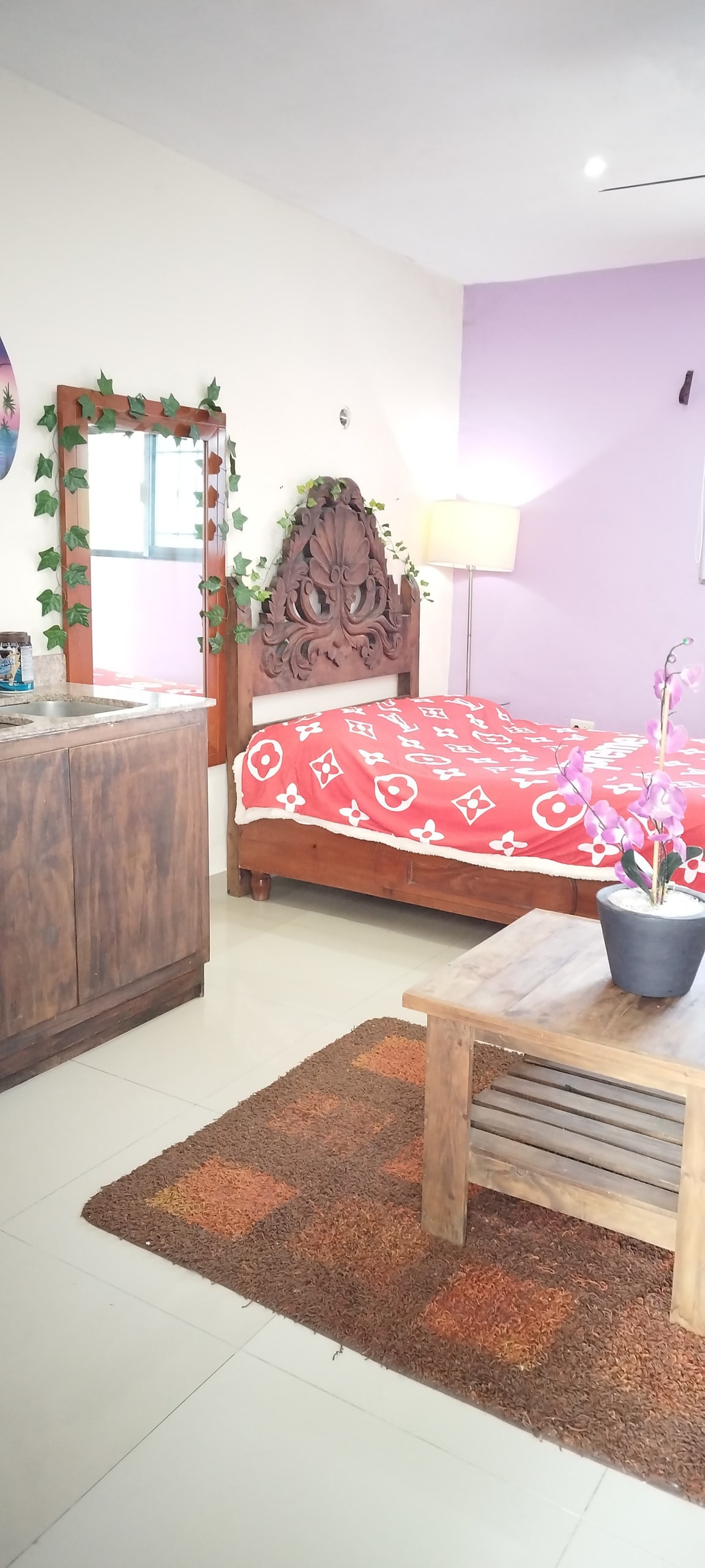 A warm bedroom features a spacious bed adorned with a patterned blanket. A decorative wooden headboard adds character. A wooden table sits in front, accompanied by a plant. A mirror reflects the space, while soft light is provided by a nearby lamp. A vibrant wall adds color.