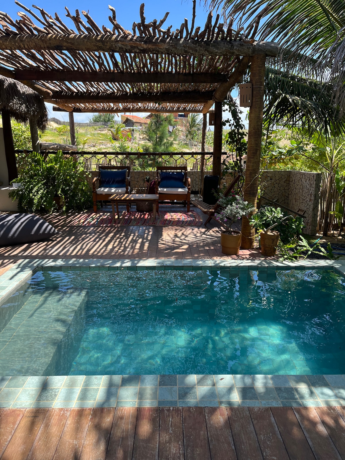 An outdoor area features a small pool surrounded by wooden decking and vibrant greenery. Two lounge chairs are positioned on a shaded terrace, offering a relaxing spot. Potted plants are neatly arranged along the edge, enhancing the natural ambiance.