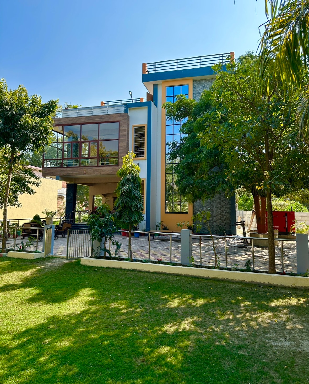 A modern three-story house is surrounded by greenery, showcasing large glass windows that reflect sunlight. A neatly maintained garden features lush grass, and the entrance is framed by tall trees, providing a sense of tranquility and connection to nature.
